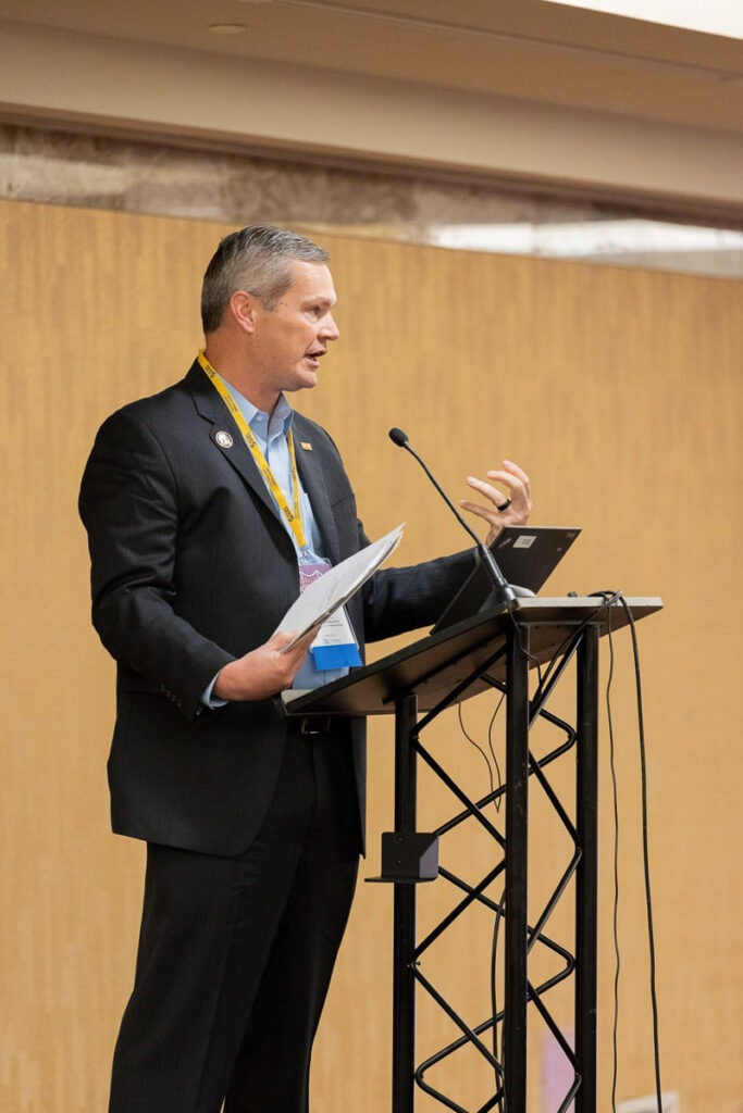 A man in business attire delivering a presentation or speech from a podium with a microphone and laptop.