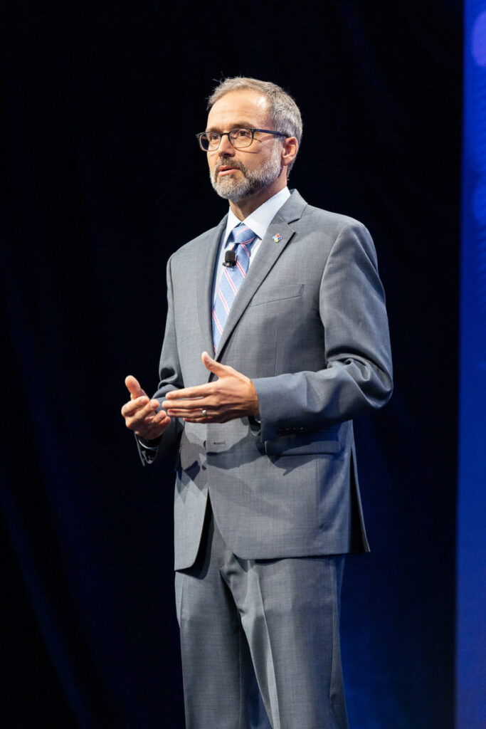 A speaker in a gray suit stands confidently on stage, addressing an attentive audience against a backdrop of soft purple lights.