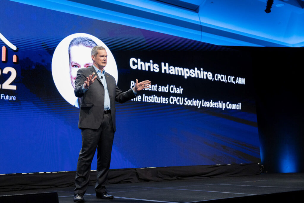 A professional speaker addressing an audience at a conference, with a presentation slide in the background featuring his name and titles.