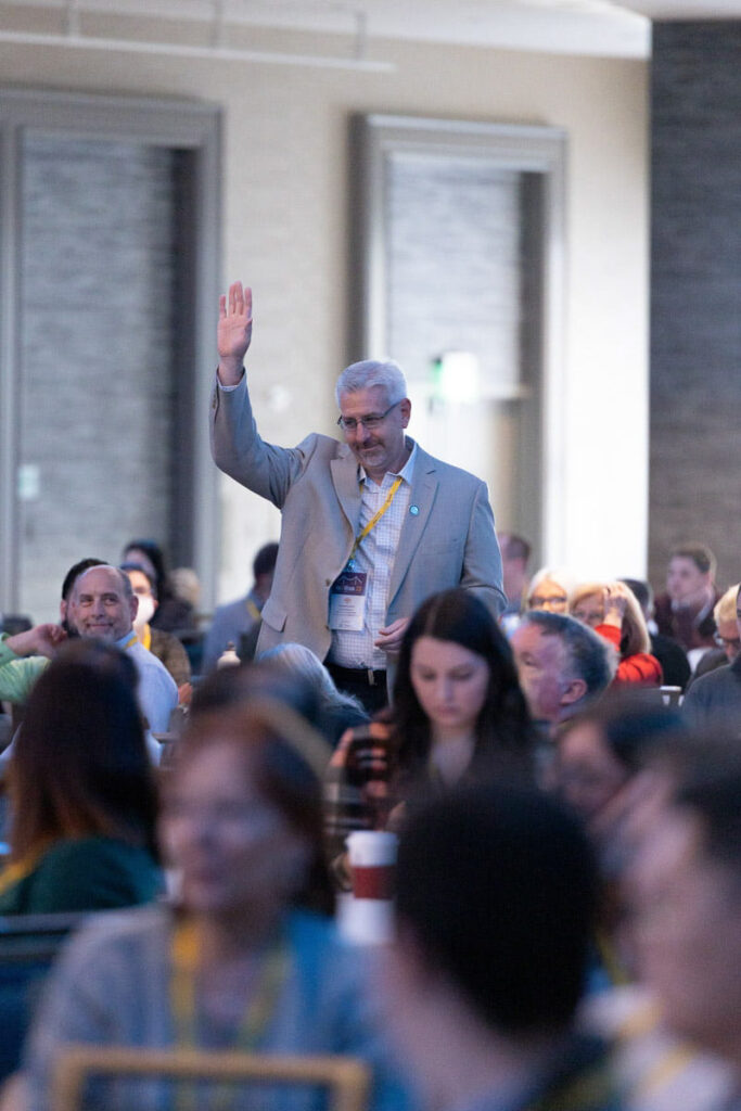 A man raising his hand to ask a question or participate during a conference session, surrounded by other attendees who are seated and focused elsewhere.