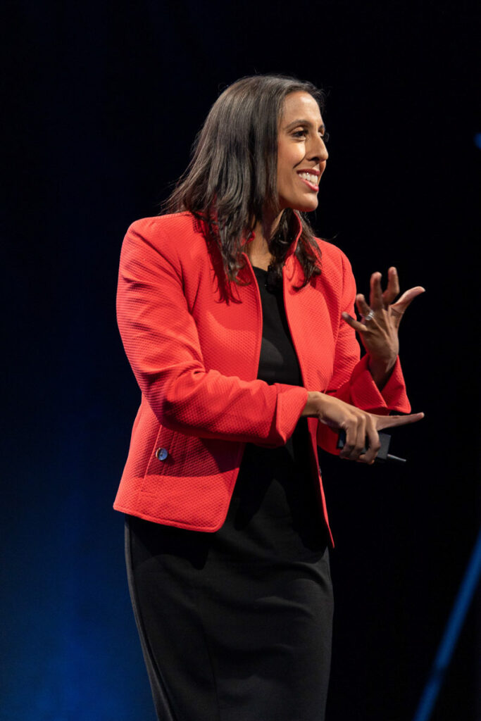 A professional woman in a vibrant red jacket gesturing with her hands while speaking at a presentation or conference.