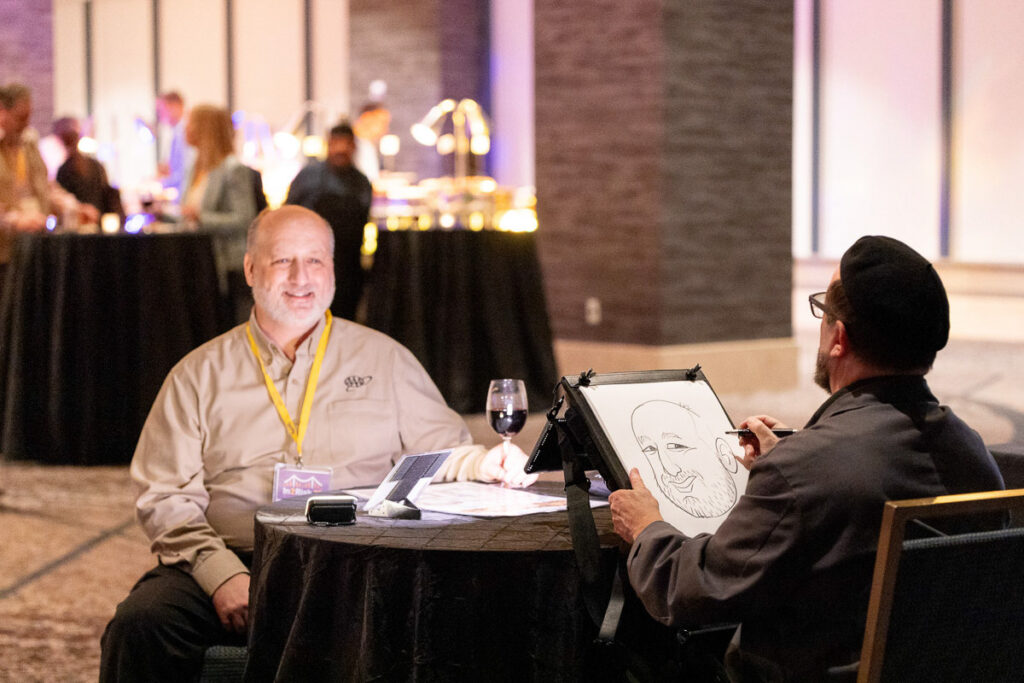 A smiling man watches as a street artist captures his likeness in a caricature drawing at an evening event.