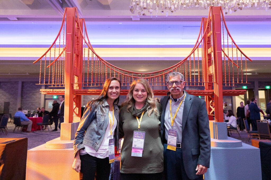 Three smiling attendees wearing conference badges stand in front of a display featuring a model of the golden gate bridge at a convention or networking event.