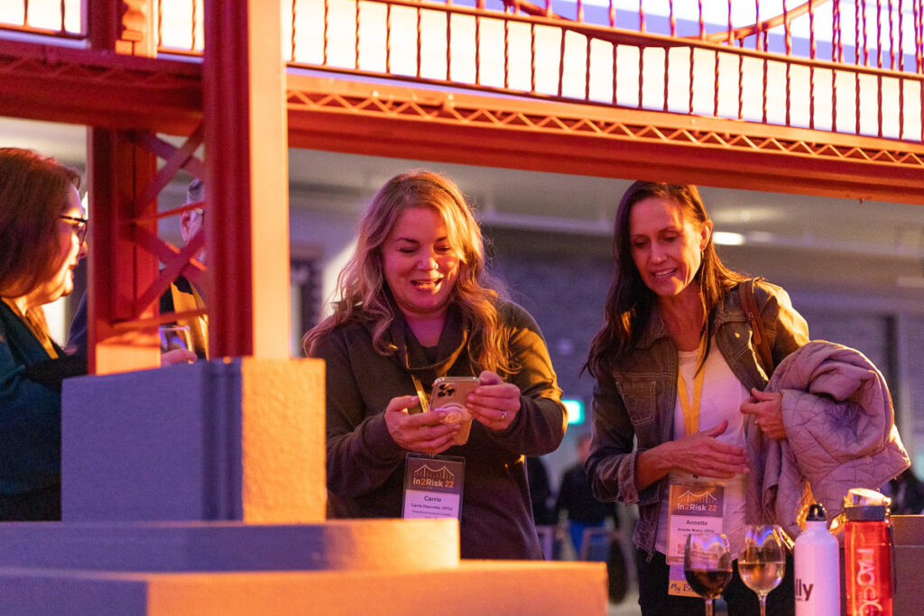 Two women sharing a joyful moment while one of them shows something on a smartphone, with warm ambient lighting creating a cozy evening atmosphere at an outdoor event.