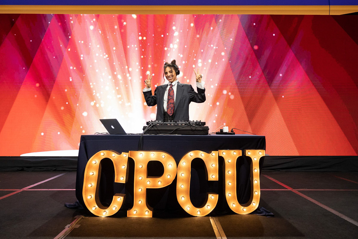 A dj with a radiant smile energizes the crowd from behind the decks at an event embellished with the initials "cpcu" in large illuminated letters, against a backdrop of dazzling red and pink lights.