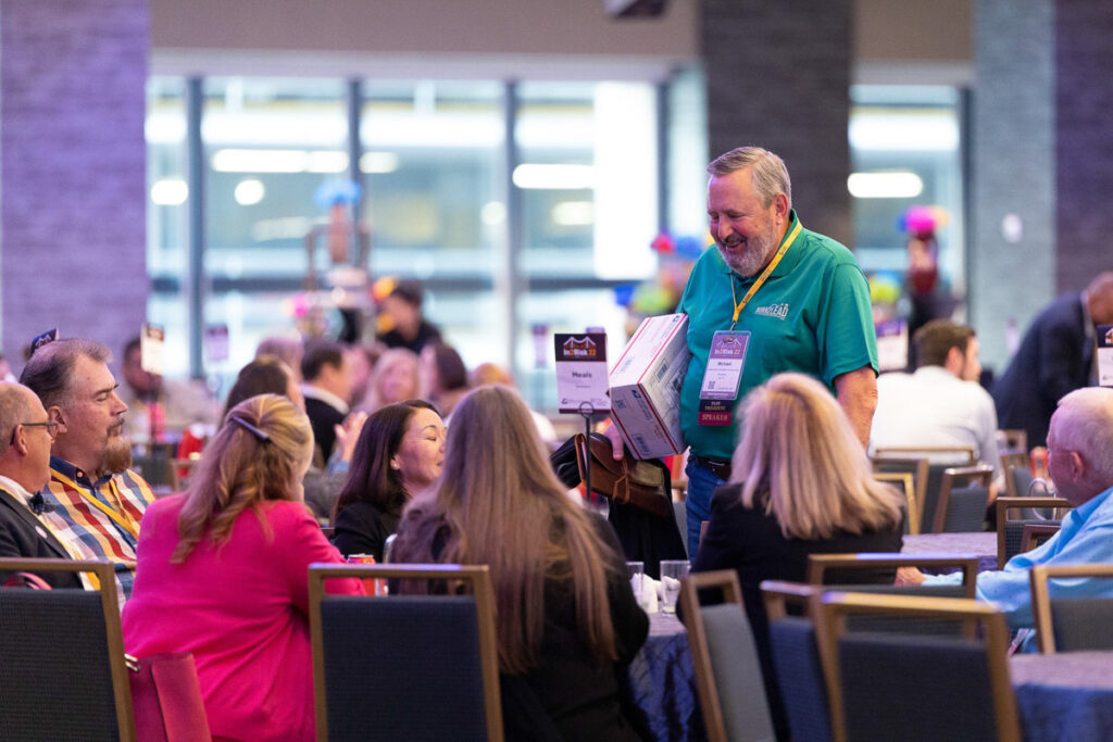 A man in a brightly colored shirt smiling as he interacts with seated attendees at a bustling indoor event space.