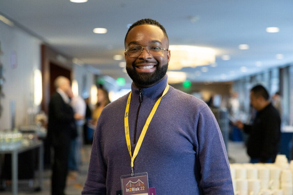 Smiling gentleman in a conference setting, wearing a purple sweater and glasses, with a badge indicating attendance at an event called "in.2.risk".