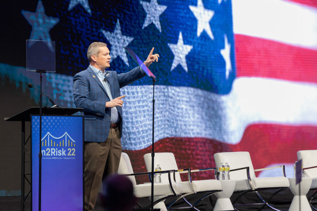 A speaker presenting at an event with a large american flag displayed on the screen behind him.