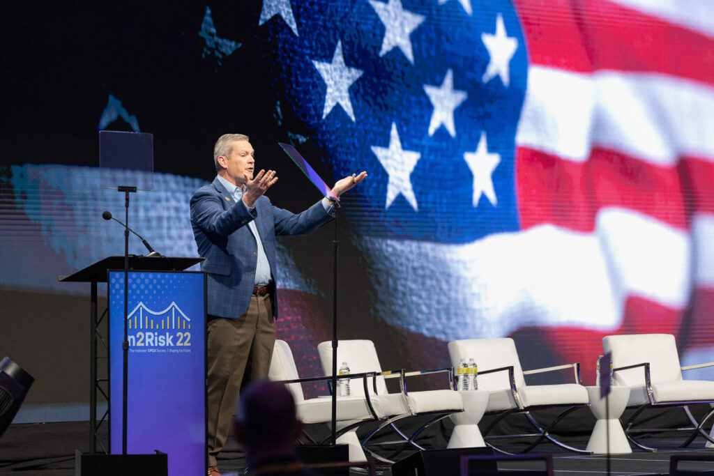 A speaker presenting at an event with a large american flag displayed on the screen behind him.