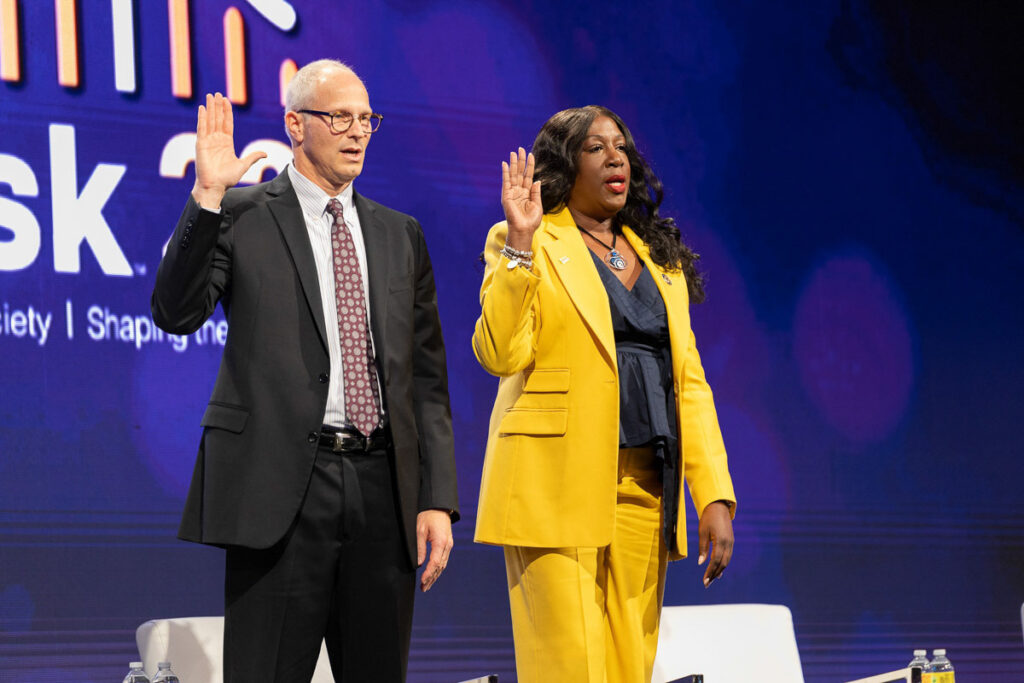 Two individuals on a stage, participating in a swearing-in ceremony against a backdrop with an event logo.