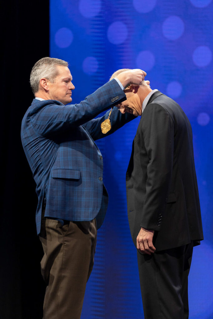 A man in a blue plaid jacket placing a medal over the head of another man in a black suit who is bowing slightly, set against a backdrop of a blue lit stage.