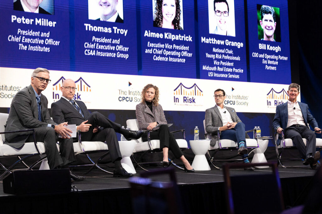 Panel of corporate professionals engaged in a discussion at a conference with their names and titles displayed on a large screen in the background.