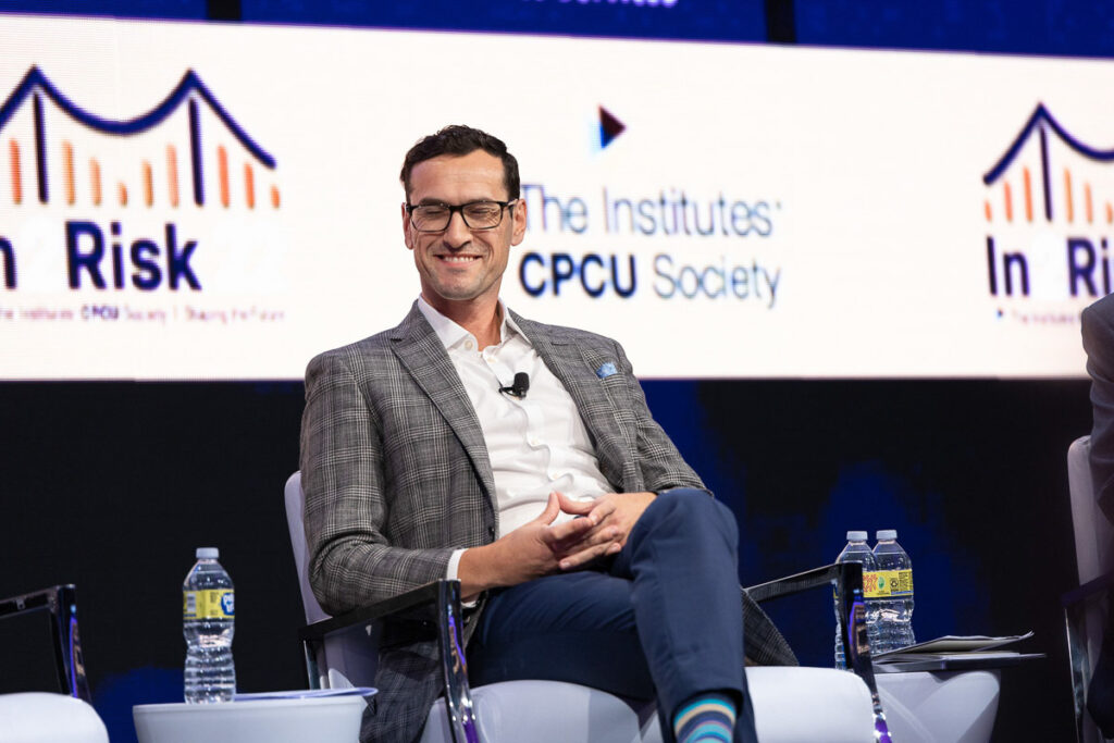 Matthew Grange sitting on stage with a microphone, participating in a panel discussion at a conference event focused on risk management, as indicated by the backdrop graphics.