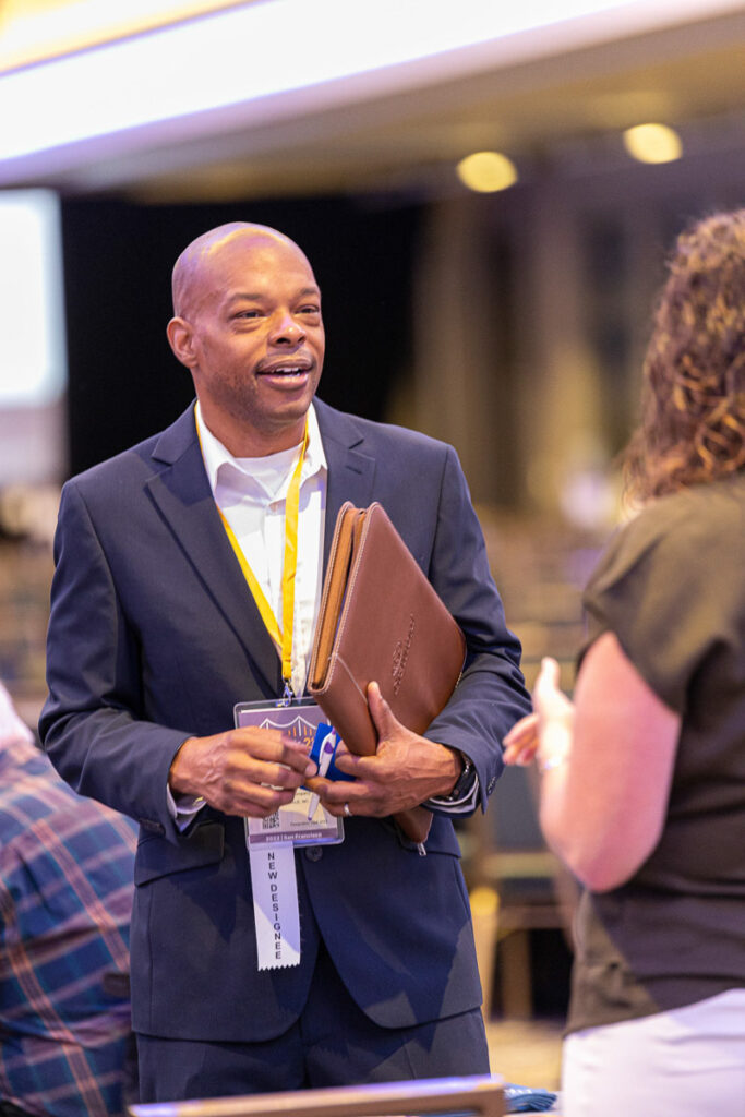 A cheerful man in business attire engaging in a lively conversation at a conference, holding a notebook and wearing a lanyard with a name tag.