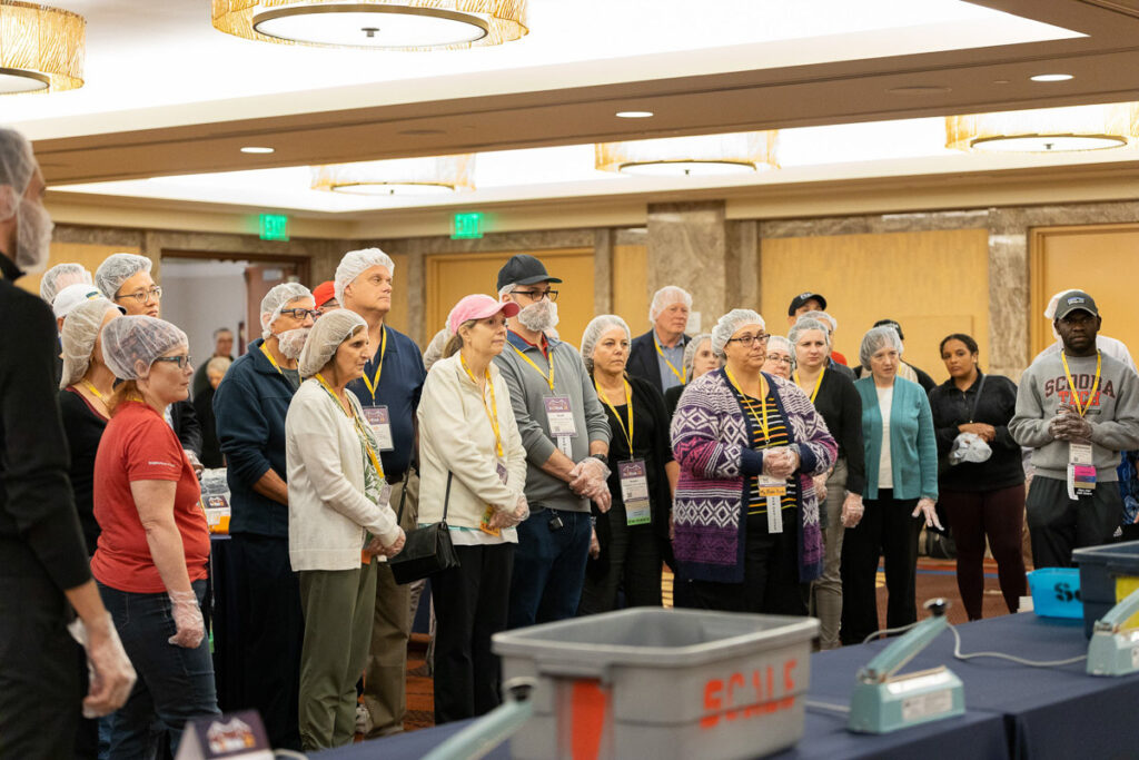 A group of volunteers attentively listening to instructions before starting a food packing activity, ready with hairnets for hygiene and name tags for identification.