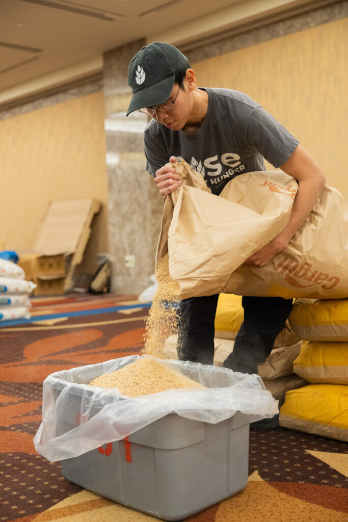 Volunteer carefully pouring a large bag of grains into a container as part of a food packing event.