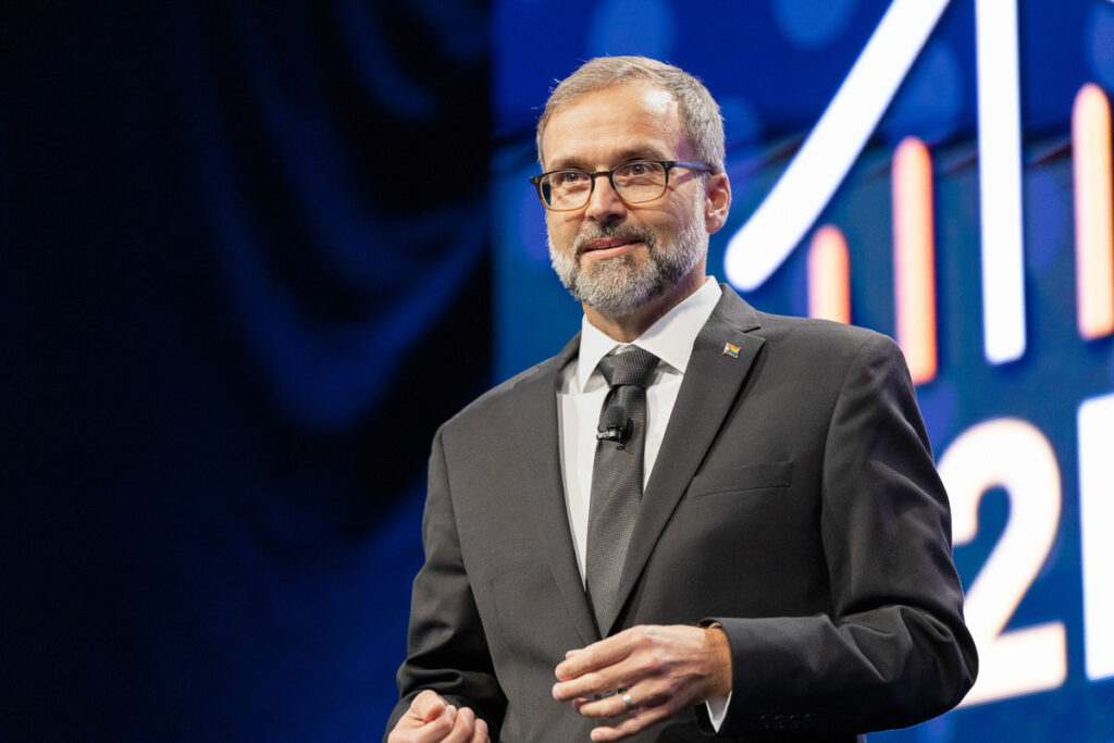 A man in a smart suit with glasses speaking at a conference, with a dynamic and colorful graphic.