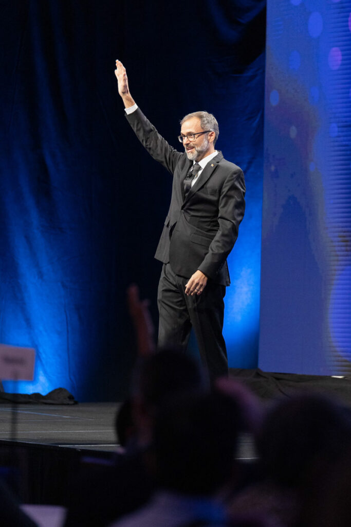 A man in a formal suit confidently raising his hand to acknowledge an audience at a photography event, possibly signaling his readiness to speak or to answer a question.