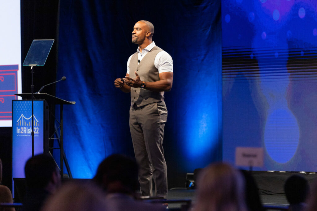 A confident keynote speaker presenting at an event with attentive audience in the foreground.