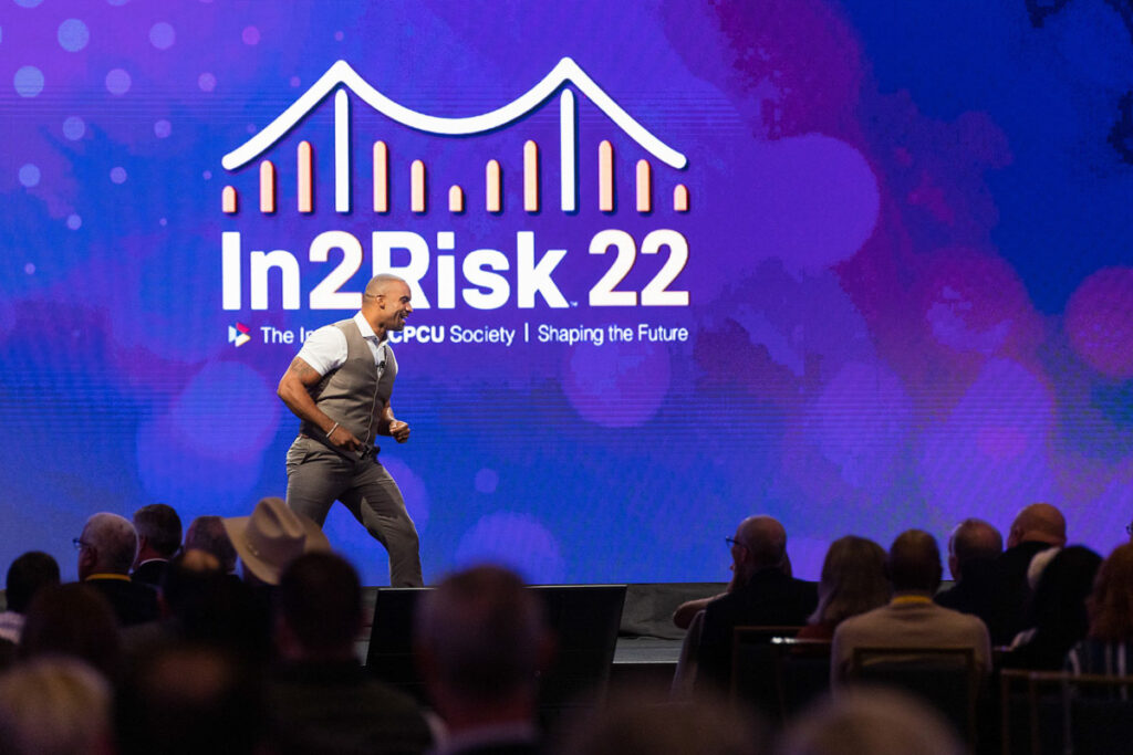 A keynote speaker confidently strides across the stage during the "in2risk 22" event under a digital screen displaying the logo and theme of the conference.