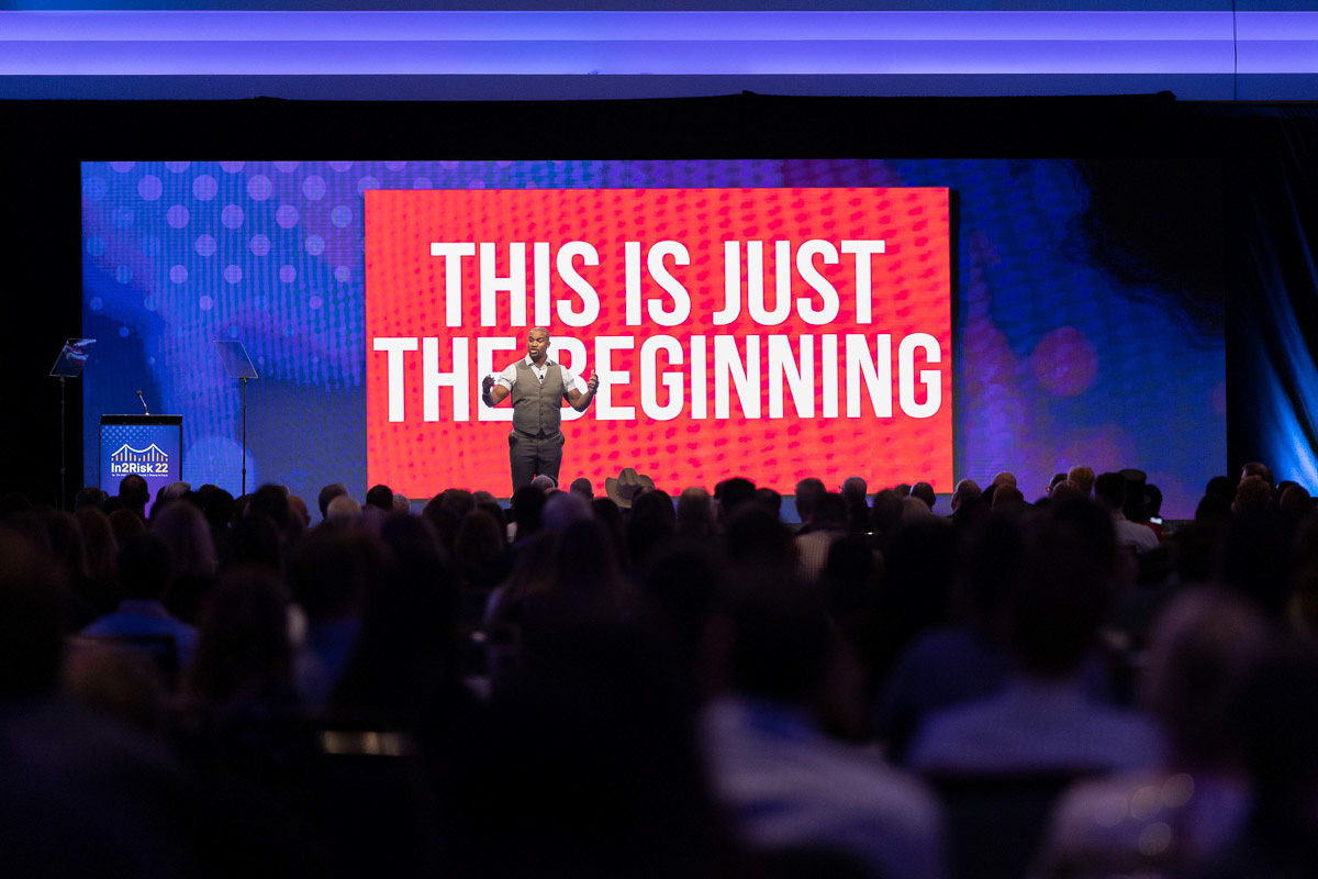 A speaker addresses an attentive audience at a conference with an inspiring message on the screen stating "this is just the beginning," captured through event photography.