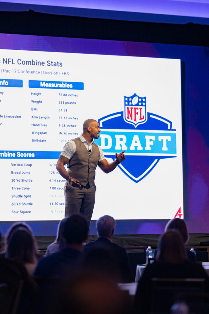 A keynote presenter speaks at an event in front of a large screen displaying NFL draft and combine stats.