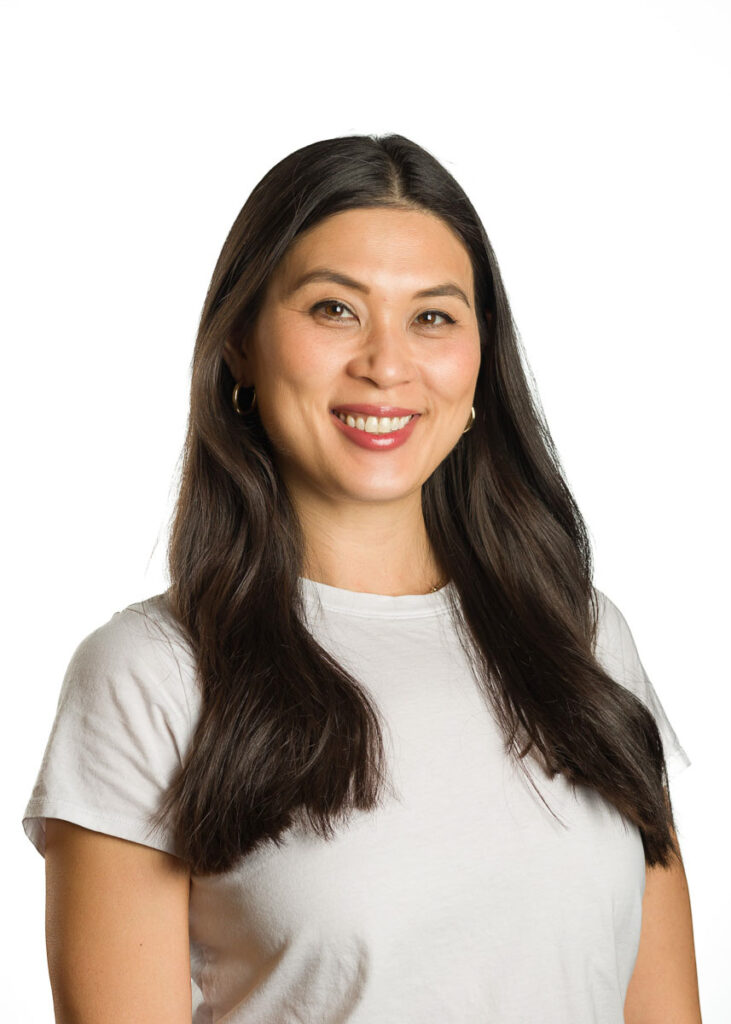 A smiling woman with long dark hair wearing a white t-shirt, posing for a corporate headshot against a white background.