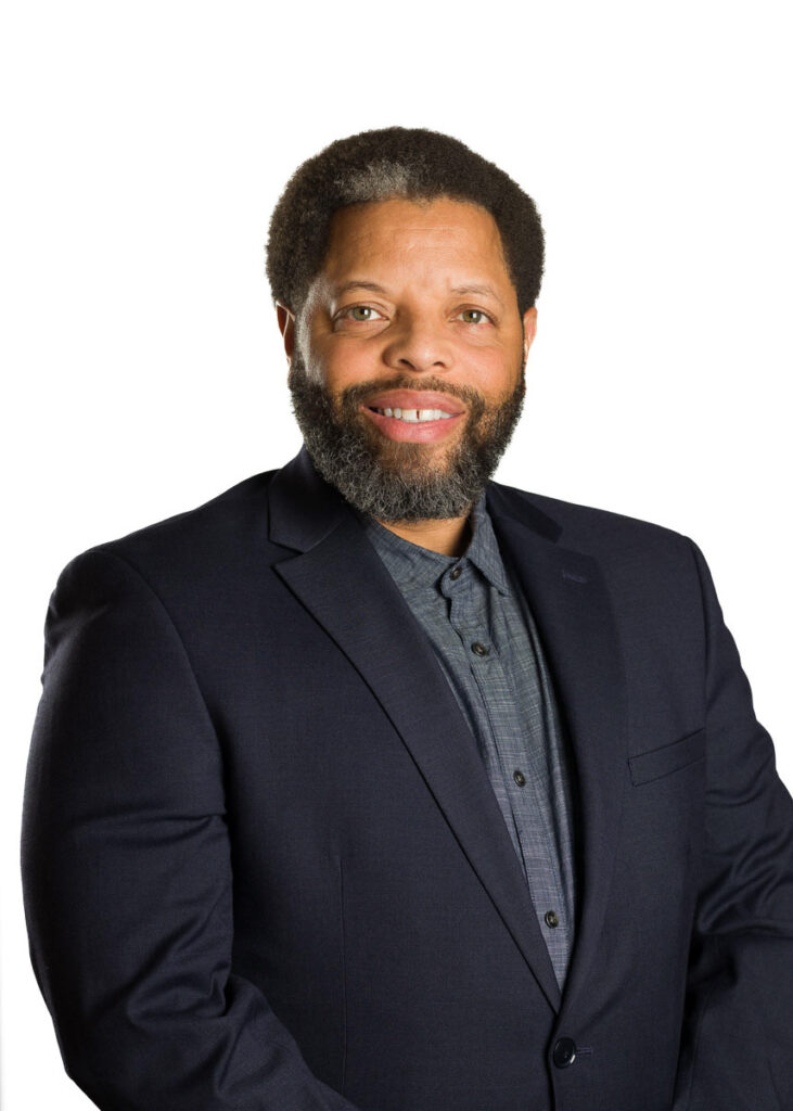 A professional man sporting a beard and a warm smile, dressed in a smart navy blue suit with a buttoned-up shirt, standing against a white background for his corporate headshot.