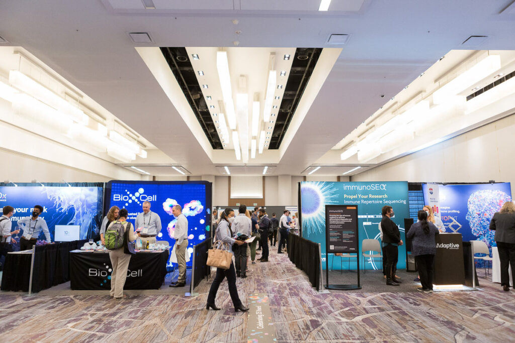 An active conference hall with attendees engaging with exhibitors at various informational booths related to biotechnology and scientific research.