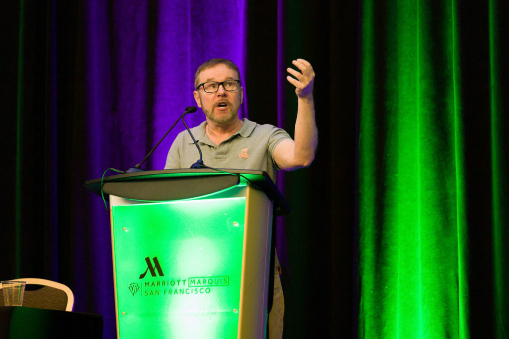 A man giving a presentation or speech at a podium labeled "Marriott marquis San Francisco" with a gesture that suggests he is emphasizing a point or asking a question.