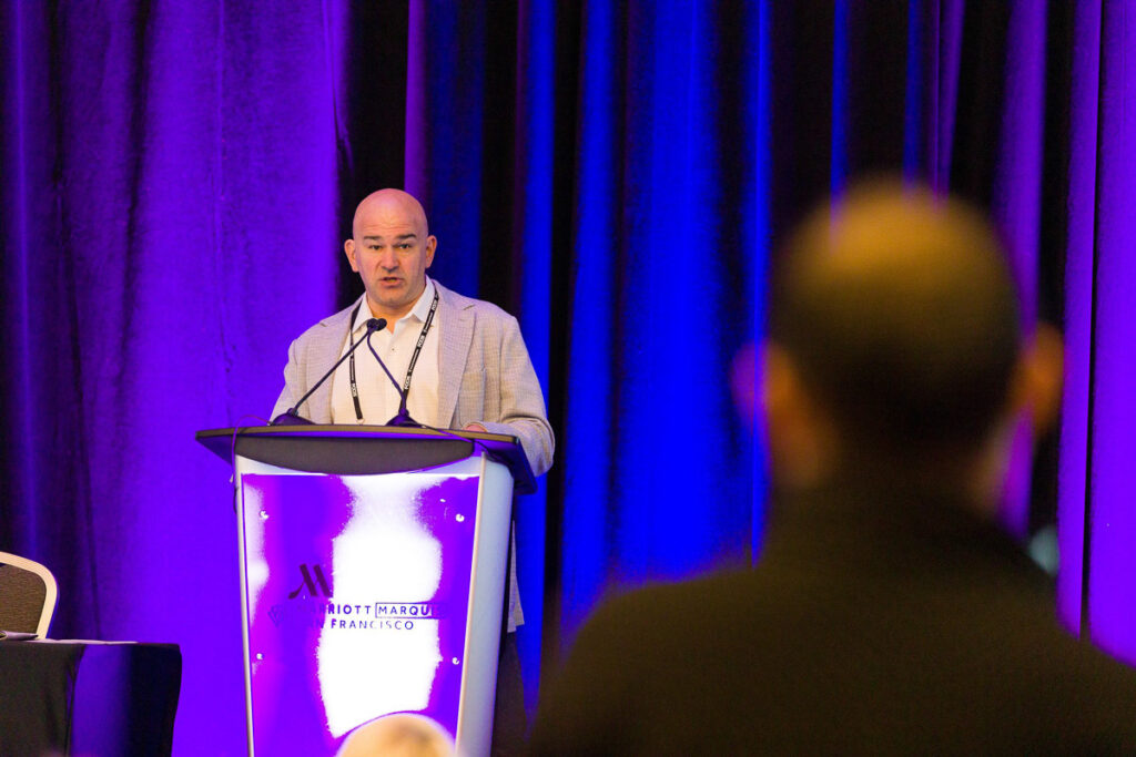 A speaker presenting at a podium with a purple and white illuminated background, possibly during a conference or seminar, with an attentive audience member in the foreground.