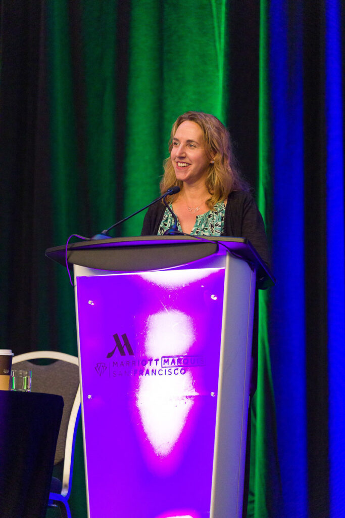 A smiling woman presenting at a podium with the Marriott marquis San Francisco logo, with a backdrop of green drapes.