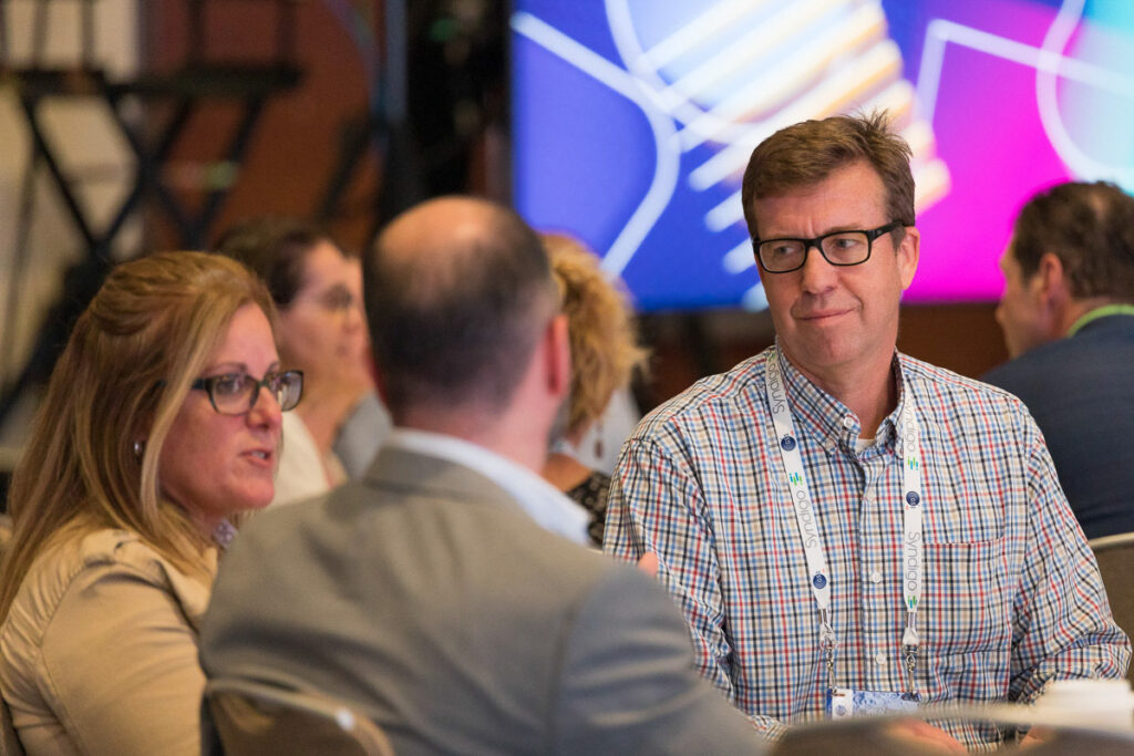 Attendees in a discussion at a conference event, with a focus on an engaged man wearing glasses and a checkered shirt looking attentively to his side.