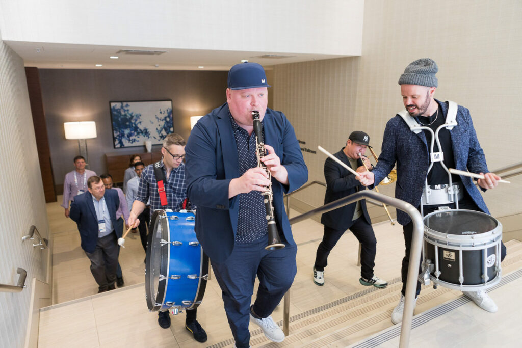 A lively impromptu band performance in a hotel corridor during a conference, featuring a saxophonist, a drummer, and a percussionist, drawing the attention of amused onlookers.