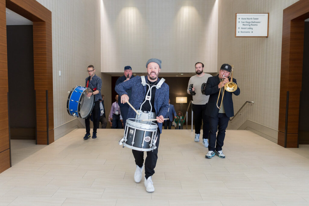 A lively band procession inside a conference building, with the lead musician playing a snare drum and marching ahead, followed by bandmates with a bass drum, trombone, and trumpet.