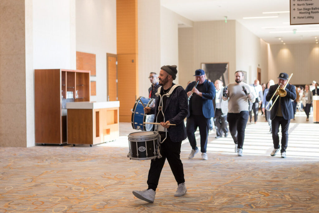 A drummer leads a procession of brass musicians through a spacious indoor hallway at a conference.