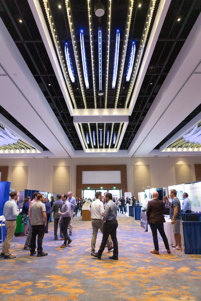 Professionals network and explore exhibit booths at a busy industry conference hall with modern overhead lighting.