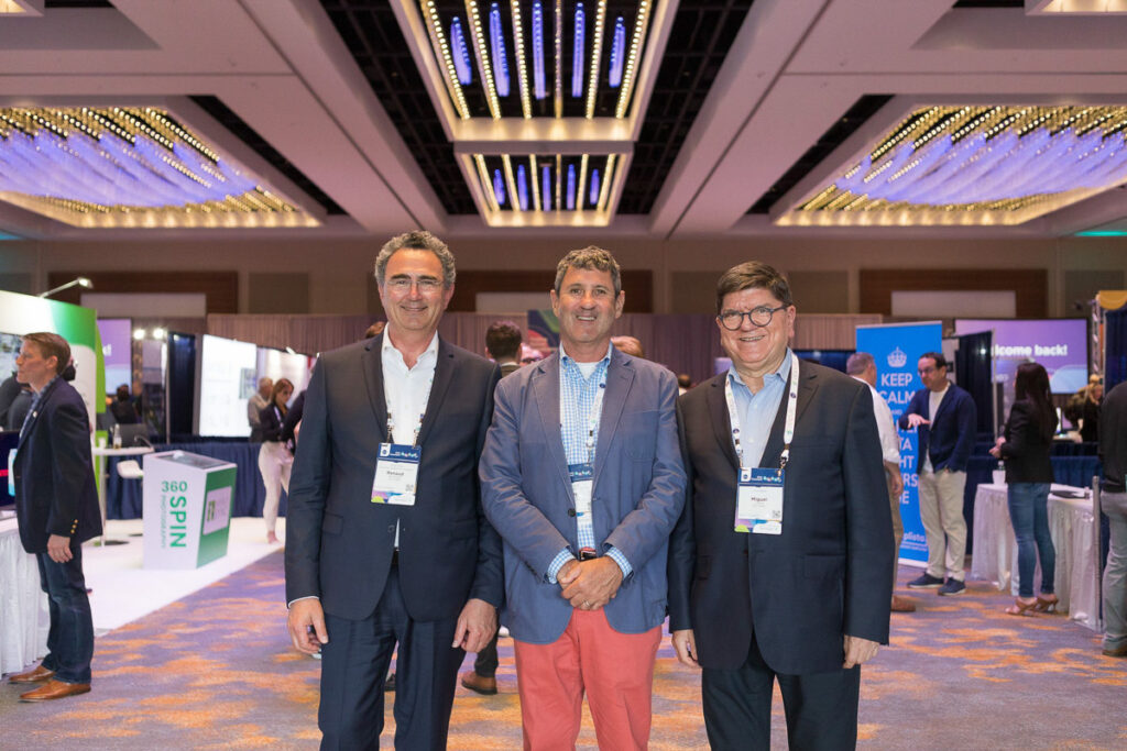 Three smiling men wearing conference badges pose together inside a convention center with exhibit booths in the background.