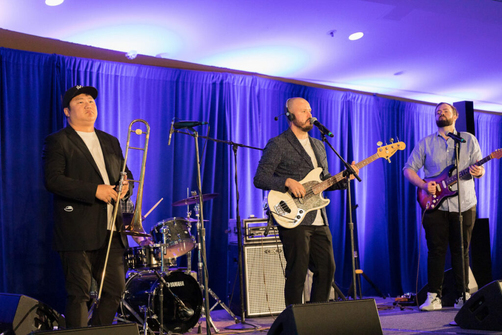 A trio of musicians performing on stage with a trombonist on the left, bass guitarist in the middle, and a singer with an electric guitar on the right, under elegant blue lighting.