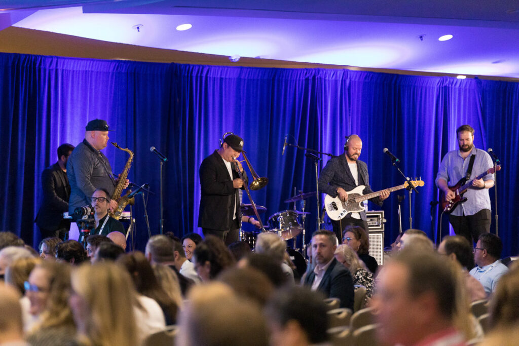 A live band performing on stage at an event with an audience seated at tables enjoying the entertainment.