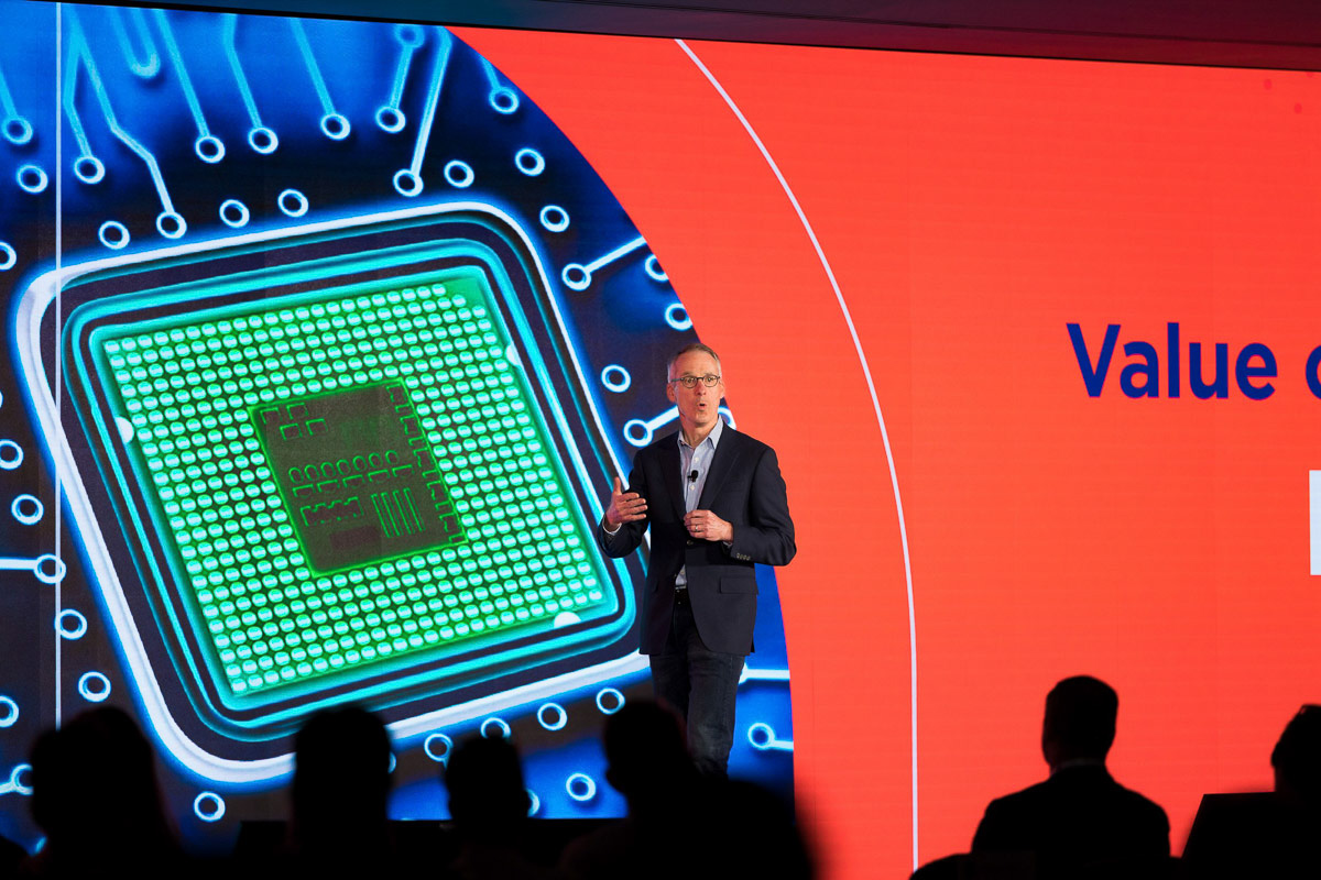 A presenter speaks at a technology conference with a giant backdrop displaying a microchip graphic.