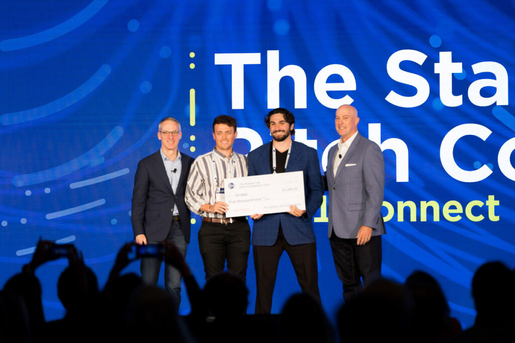 Four individuals on a stage at a health conference, with the central figure holding a large ceremonial check, possibly signifying a grant or prize award, while others applaud or pose for pictures.