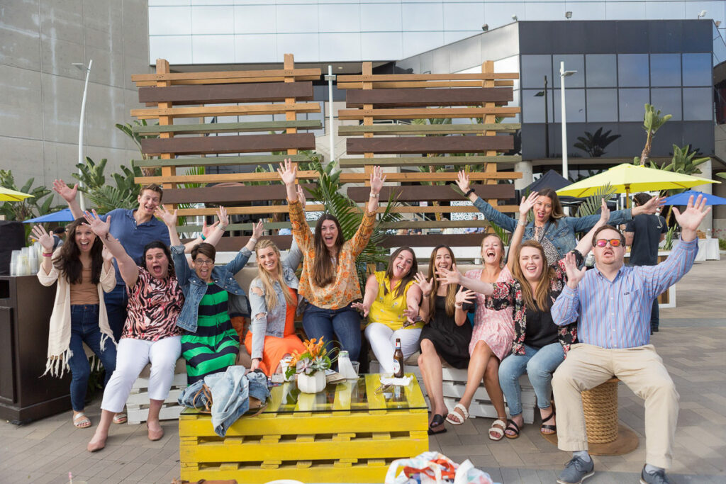 A joyful group of people posing for a photo with wide smiles and raised arms, exuding happiness and excitement at an outdoor conference gathering.