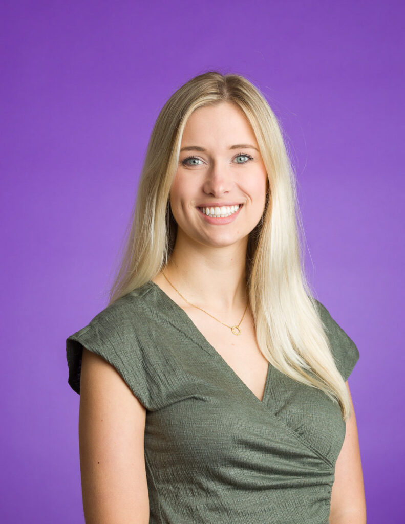 A smiling woman with long blonde hair, wearing a green top, against a purple background in a corporate headshot.