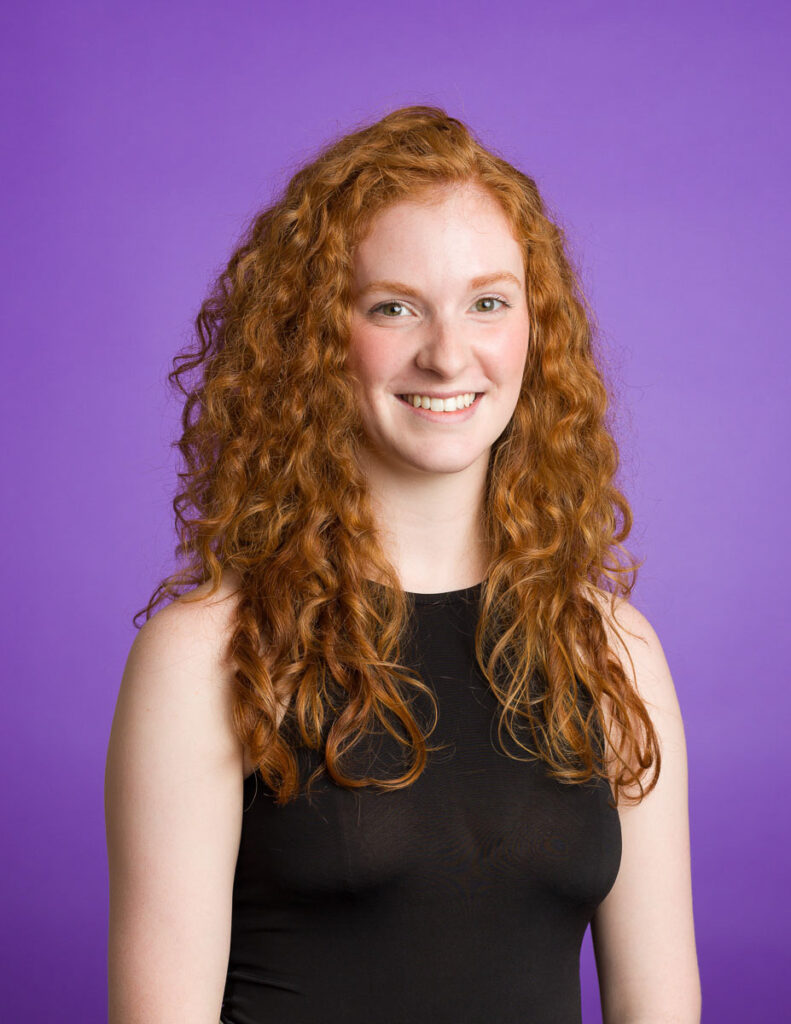 Portrait photography of a smiling woman with curly red hair against a purple background.