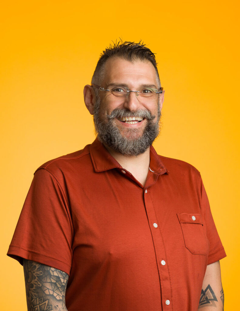 Portrait of a smiling bearded man with glasses and tattoos against a vibrant orange background.