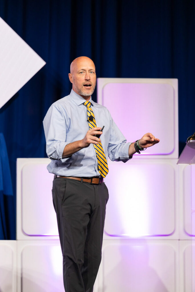 A keynote speaker presenting at a conference, gesturing with one hand while holding a remote in the other.