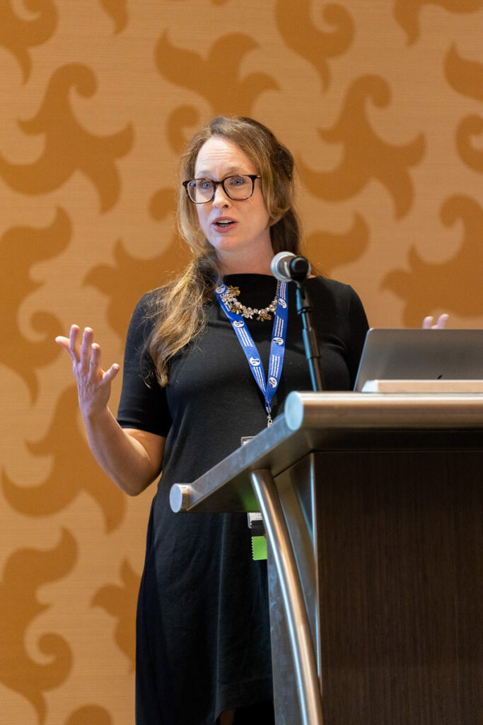 A professional woman giving a presentation at a conference, with expressive hand gestures, standing behind a lectern.