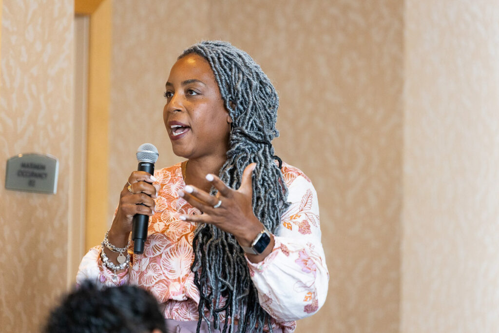 A woman with long braided hair speaks passionately into a microphone, gesturing with her hand, likely engaging an audience during a conference presentation or discussion.