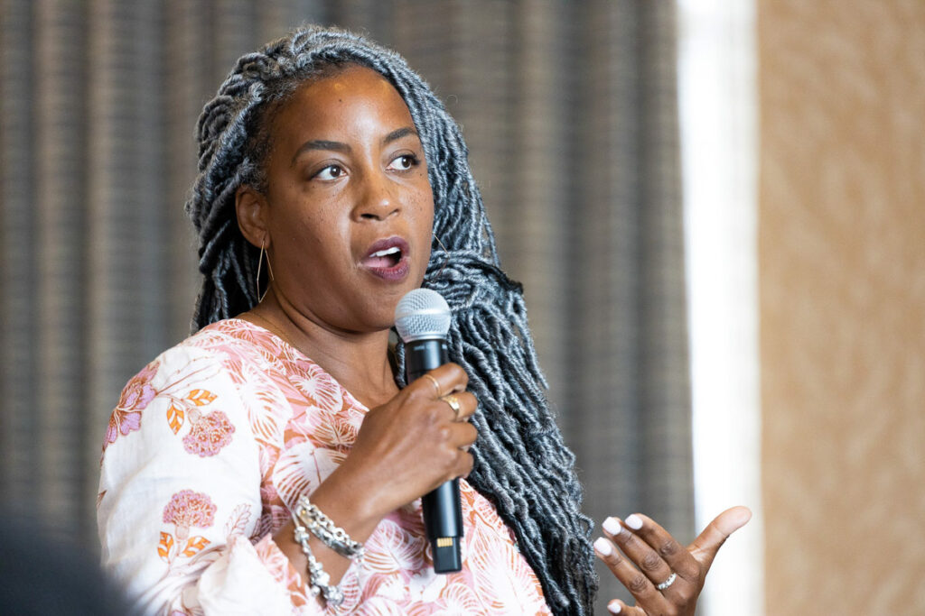 A confident speaker with long braided hair holds a microphone and gestures while speaking at an indoor conference.